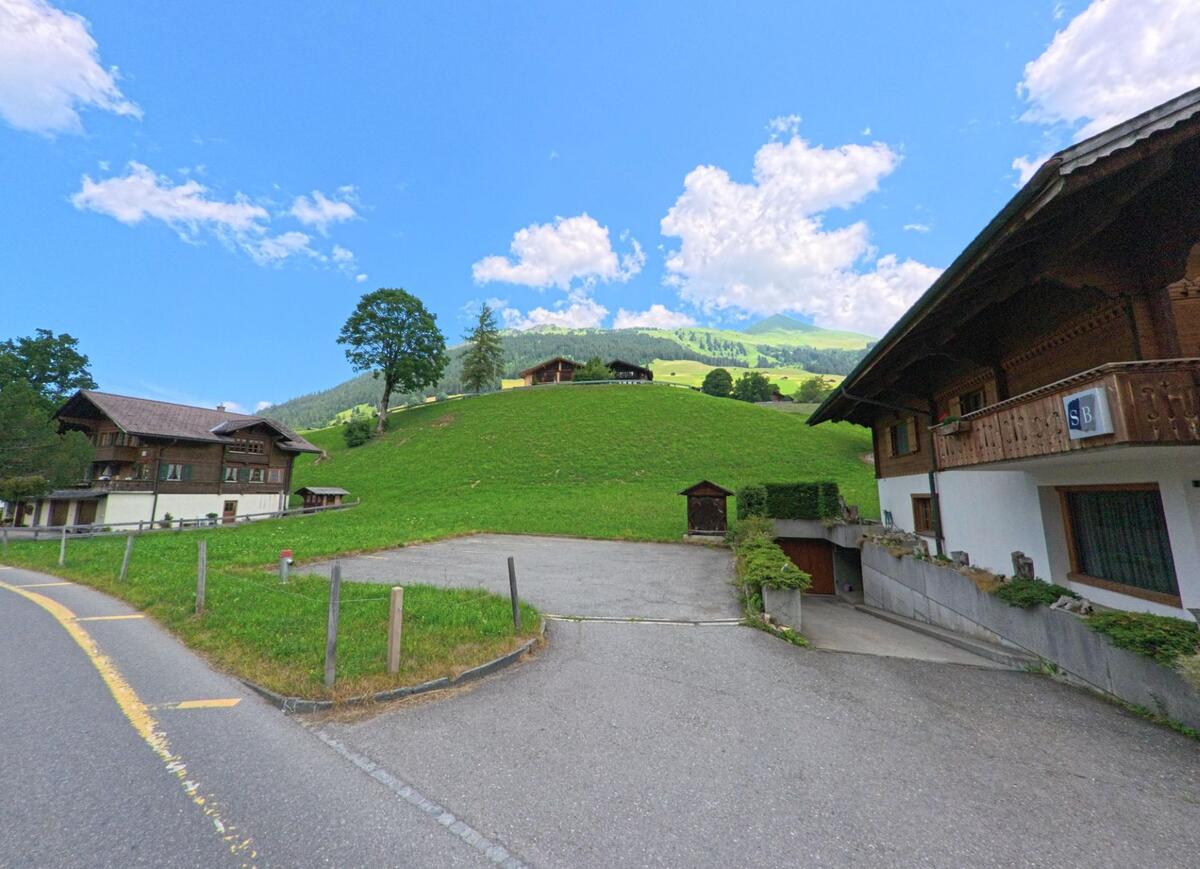 In Lauenen (3782, Kanton Bern) führt eine schmale Strasse an Wohnhäusern vorbei hinauf zu einem grasbewachsenen Hügel, auf dessen Kuppe ein Chalet unter Bäumen steht.