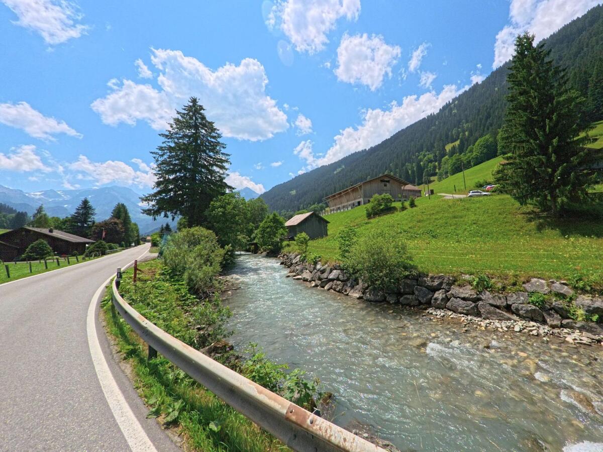In Lauenen (3782, Kanton Bern) verläuft eine Landstrasse entlang eines klaren Gebirgsbaches, flankiert von Wiesen, Bäumen und vereinzelten Häusern mit Blick auf die Alpen.