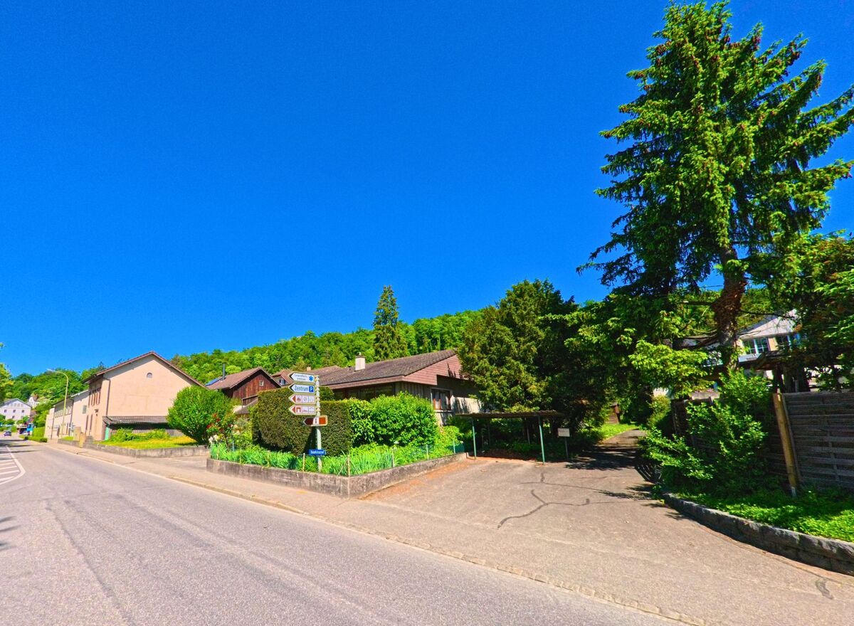 An der Strasse in Laufen (4242, Kanton Baselland) führt eine Abzweigung zu einem Wohngebiet, das von Bäumen und grünen Sträuchern umgeben ist.