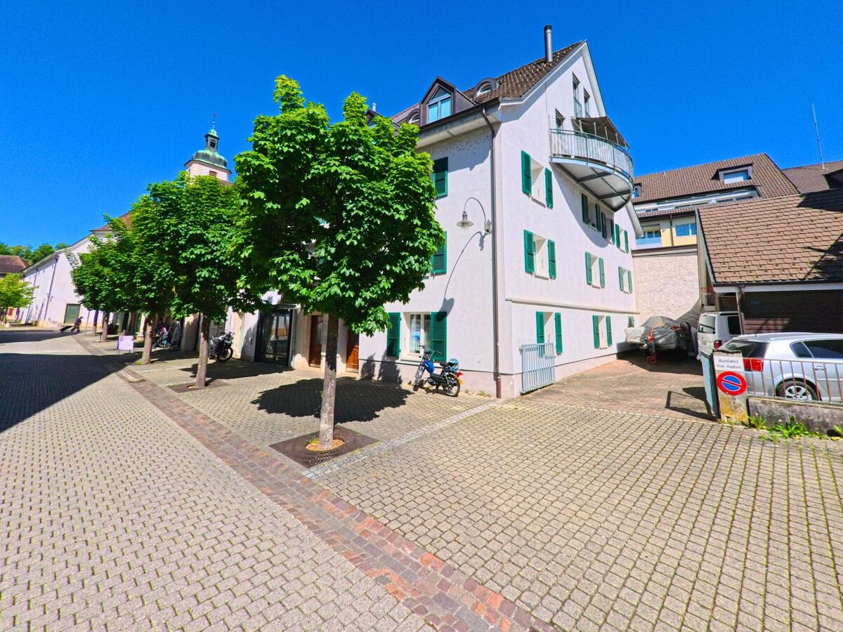In Laufen (4242, Kanton Baselland) steht ein helles Wohnhaus mit gruenen Fensterlaeden und Balkon an einer gepflasterten Allee mit regelmaessig gepflanzten Baeumen.