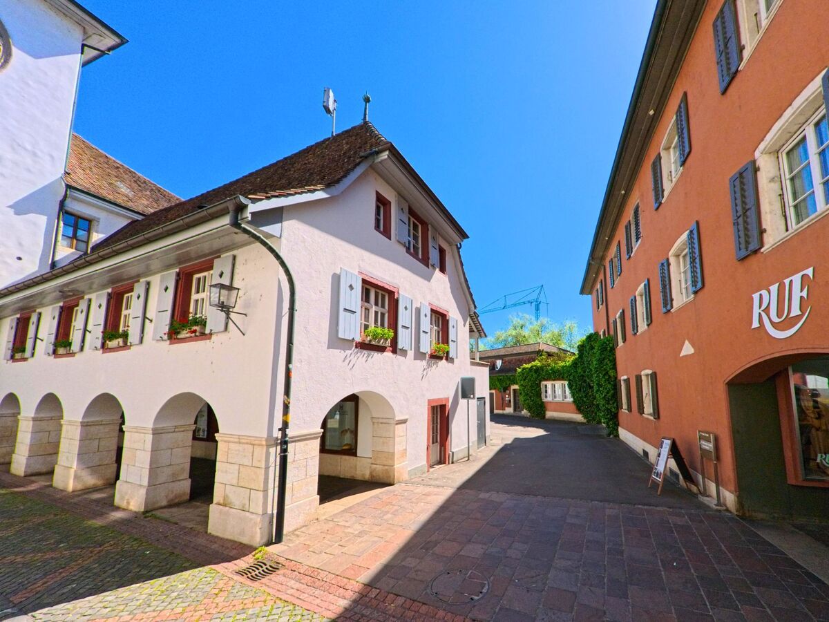 In einer gepflasterten Gasse von Laufen (4242, Kanton Baselland) stehen historische Gebaeude mit Laubengang und Fensterlaeden im Schatten eines Innenhofs.