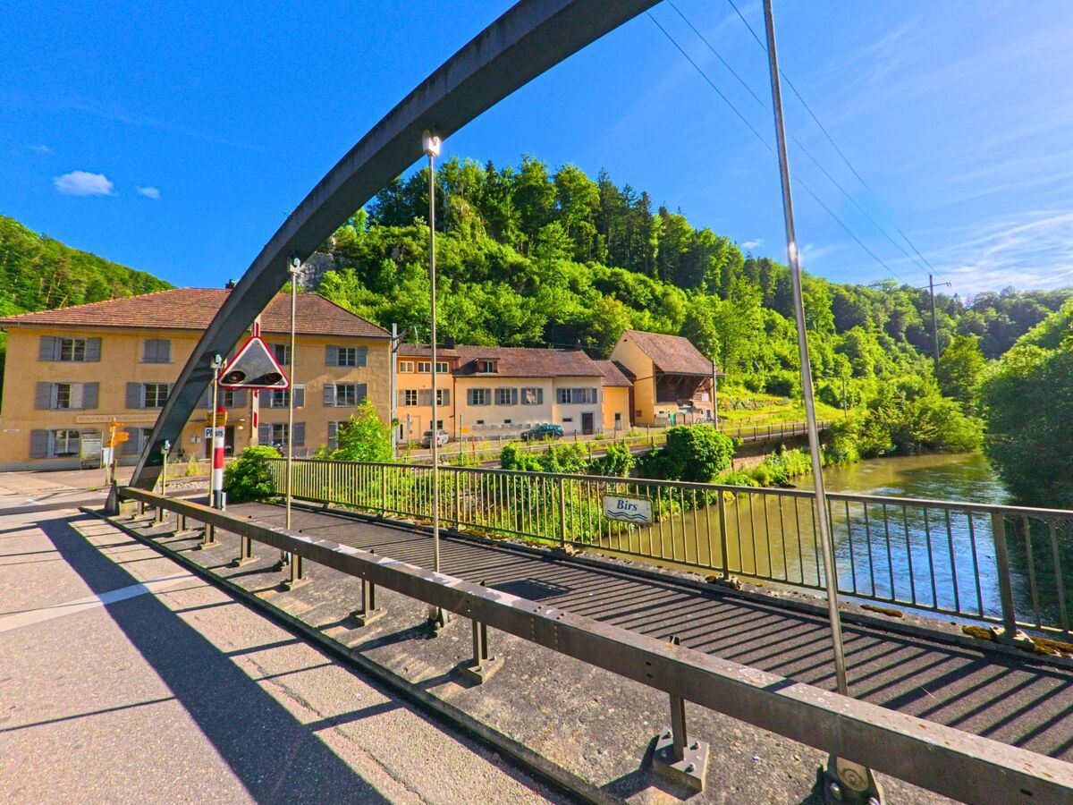 Eine Bogenbruecke fuehrt in Laufen (4242, Kanton Baselland) ueber die Birs zu historischen Gebaeuden am Flussufer, umgeben von dichter Waldlandschaft.