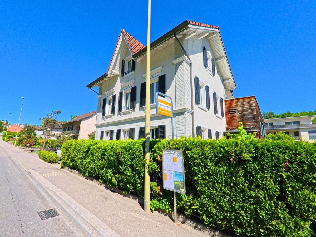 In Laufen (4242, Kanton Baselland) befindet sich eine Bushaltestelle direkt vor einem hellen Wohnhaus mit Fensterlaeden an einer sonnigen Strasse mit Baumbestand.
