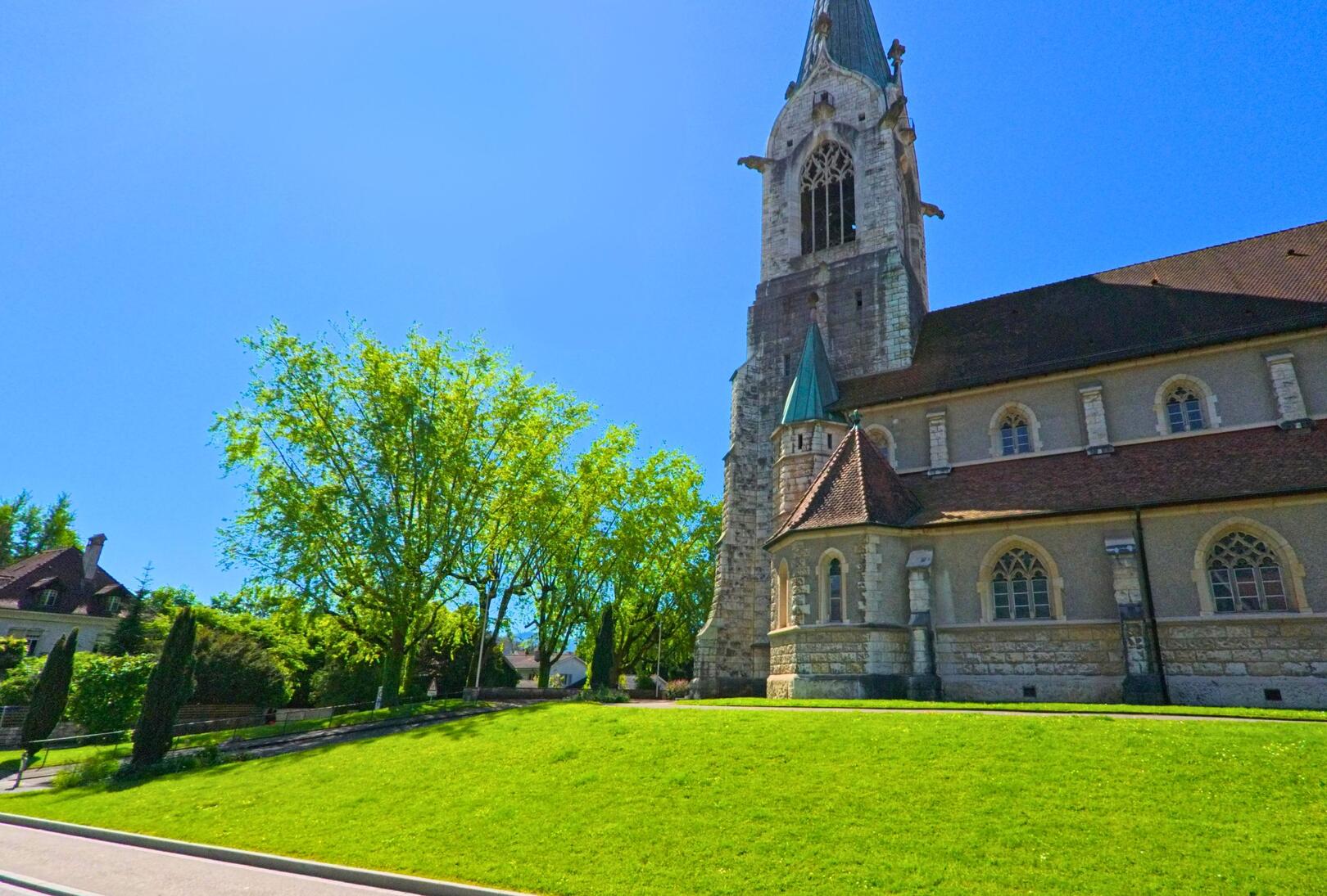 In Laufen (4242, Kanton Baselland) steht eine imposante Kirche mit neugotischem Turm auf einer leicht erhoehten Gruenflaeche direkt an der Strasse.
