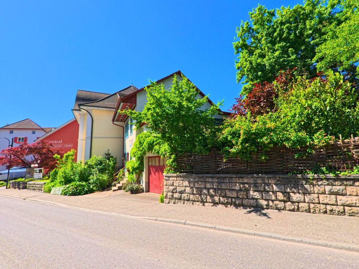 In Laufen (4242, Kanton Baselland) liegt ein begruentes Wohnhaus mit rotem Tor neben einem Hotelgebäude an einer leicht ansteigenden Strasse mit Stützmauer.