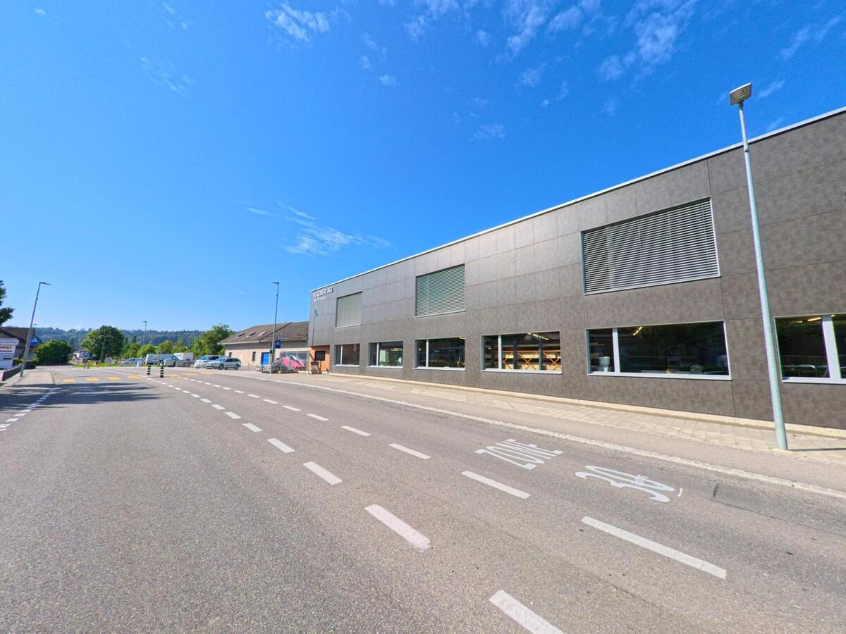 Entlang einer breiten Strasse steht ein langgezogenes Gewerbegebaeude mit grossen Fenstern in Laupen (3177, Kanton Bern) bei klarem Himmel.
