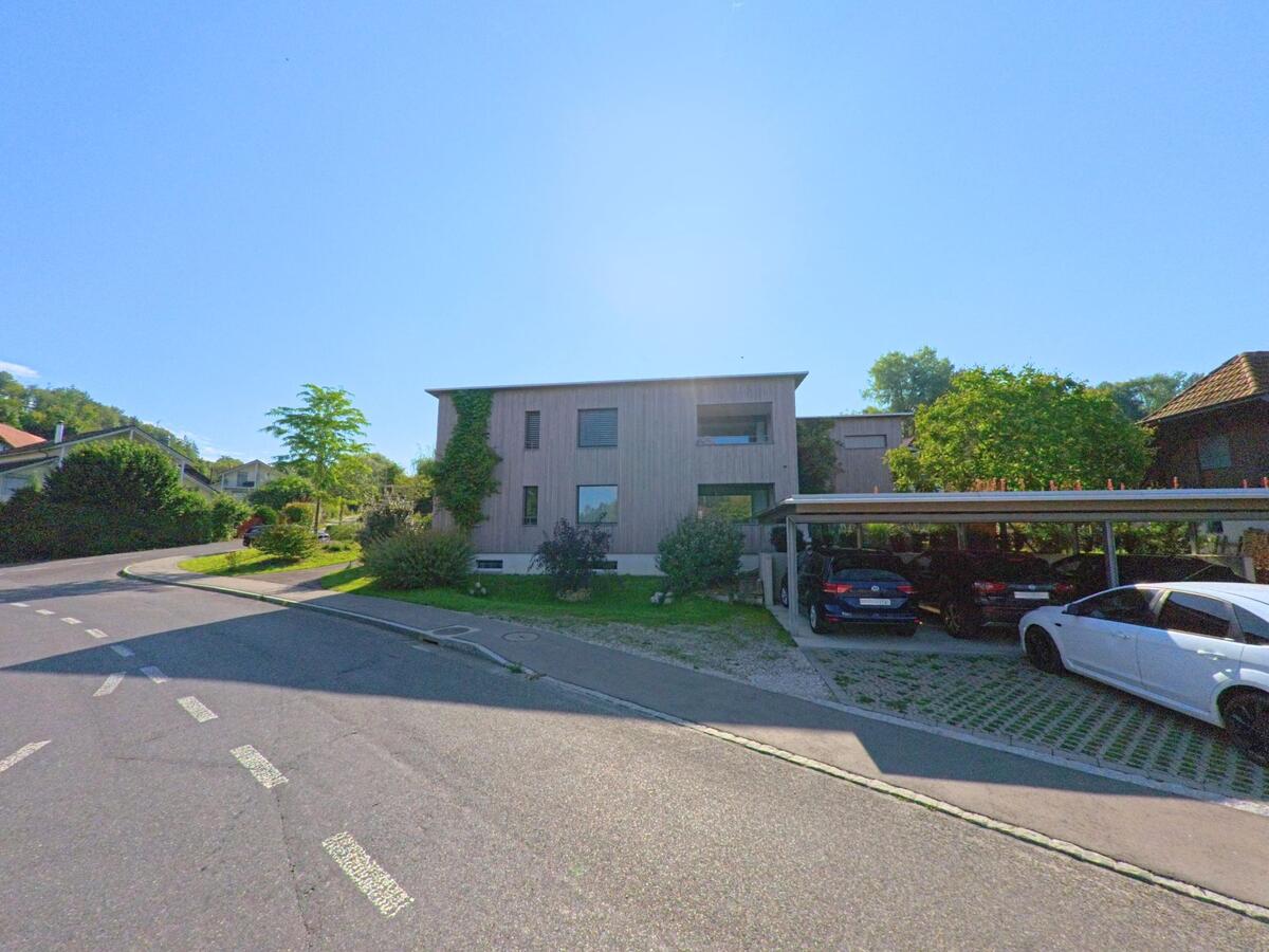 Ein modernes Wohnhaus mit Carport liegt an einer leicht ansteigenden Strasse in Laupen (3177, Kanton Bern) unter klarem Himmel.