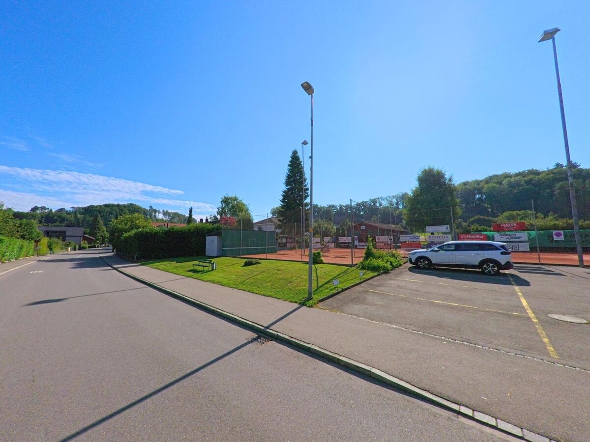 Neben einer Strasse liegt eine Tennisanlage mit Spielfeldern und Parkplatz in Laupen (3177, Kanton Bern) bei sonnigem Wetter.