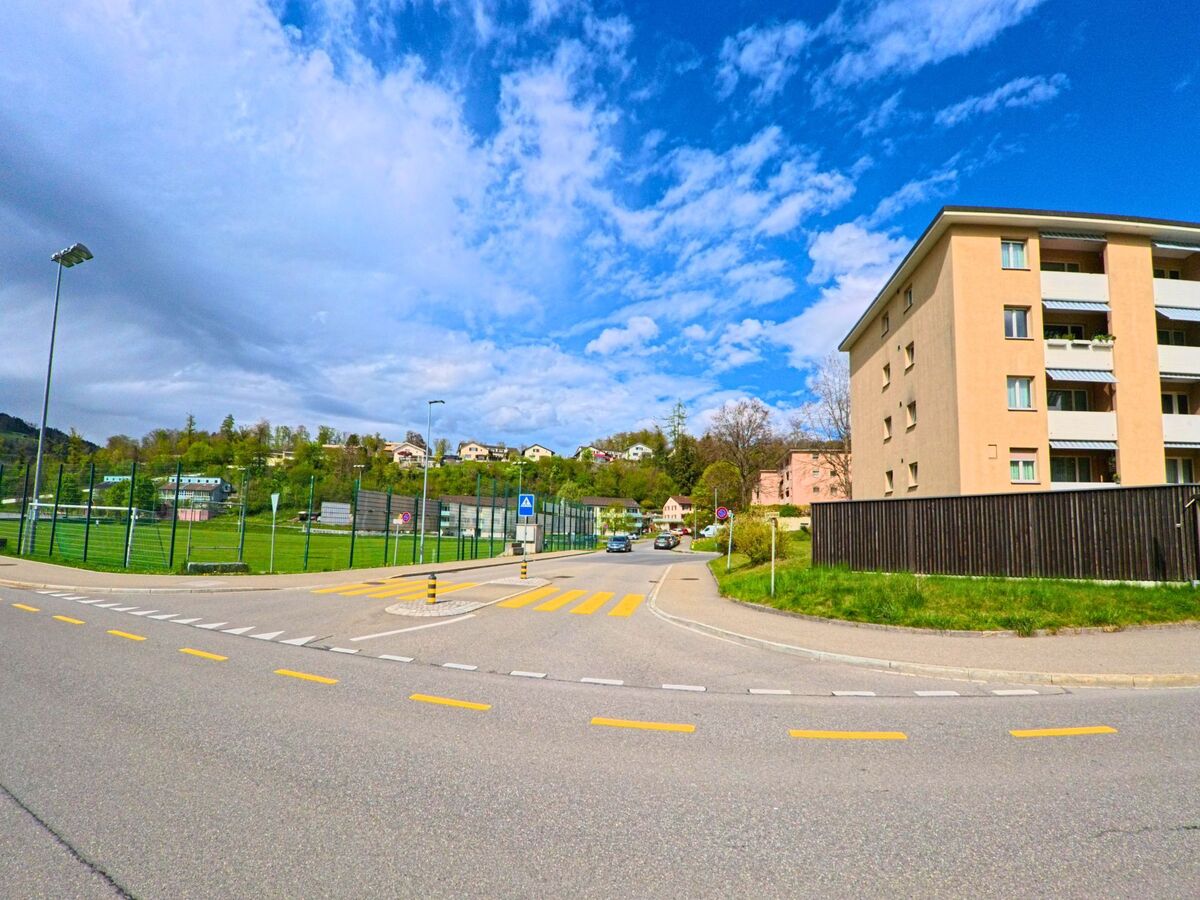 Gegenüber einem eingezäunten Sportplatz in Laupen (8636, Kanton Bern) stehen moderne Mehrfamilienhäuser entlang einer verkehrsberuhigten Strasse mit Fussgängerstreifen.