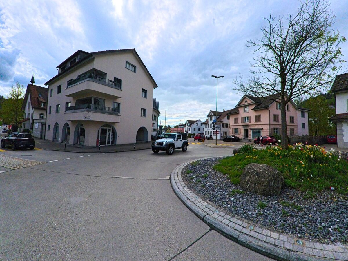 Eine begrünte Verkehrsinsel mit Frühlingsblumen liegt im Zentrum von Laupen (8636, Kanton Bern), umgeben von Wohnhäusern und einem kleinen Kreisverkehr.