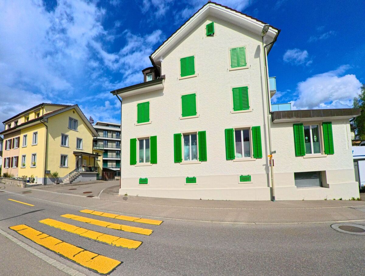Ein Zebrastreifen führt in Laupen (8636, Kanton Bern) an bunt gestrichenen Wohnhäusern mit grünen Fensterläden vorbei, unter klarem Himmel.