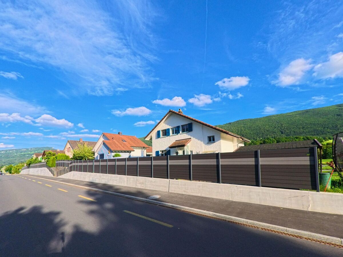 Entlang einer frisch asphaltierten Strasse in Laupersdorf (4712, Kanton Solothurn) stehen Einfamilienhäuser mit gepflegten Vorgärten und Blick auf den bewaldeten Hang.