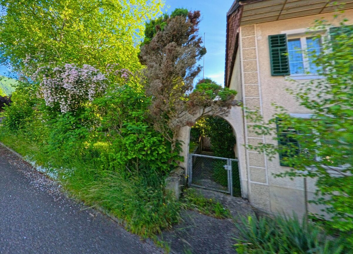 In Laupersdorf (4712, Kanton Solothurn) führt ein verwachsener Gartendurchgang mit Rundbogen und Metalltor zu einem üppig bewachsenen Grundstück neben einem Wohnhaus mit grünen Fensterläden.