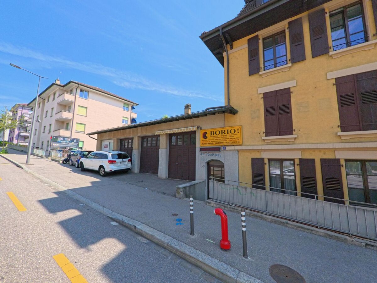 In Lausanne (1010, Kanton Waadt) steht ein gelbes Wohn- und Geschaeftshaus mit geschlossenen Fensterlaeden neben einer Strasse mit geparkten Fahrzeugen.