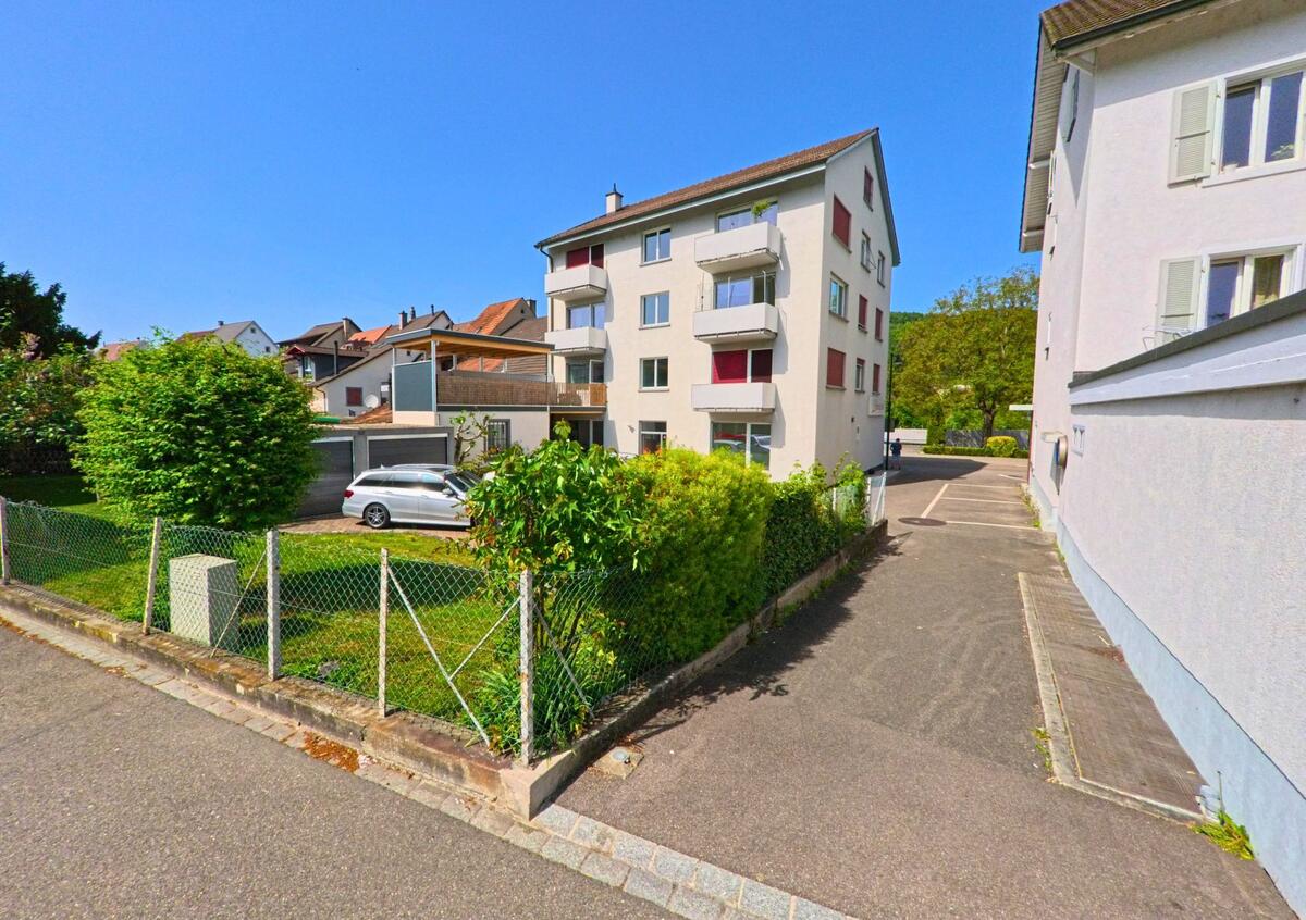 In Lausen (4415, Kanton Baselland) sind zwei Wohngebäude zu sehen, die von einem Garten und einem Parkplatz umgeben sind. Ein Auto steht auf dem Parkplatz.