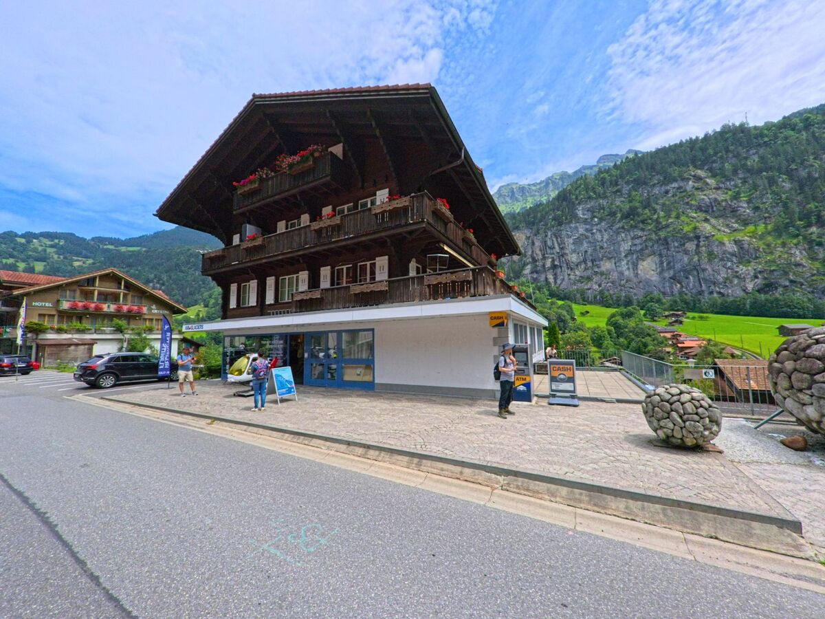 In Lauterbrunnen (3822, Kanton Bern) befindet sich ein traditionelles Chalet mit Sportgeschaeft und Bankautomat, dahinter erhebt sich eine steile Felswand vor gruener Landschaft.
