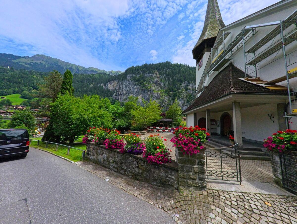Vor der reformierten Kirche in Lauterbrunnen (3822, Kanton Bern) blühen farbenfrohe Blumen, im Hintergrund erhebt sich eine bewaldete Bergkulisse.