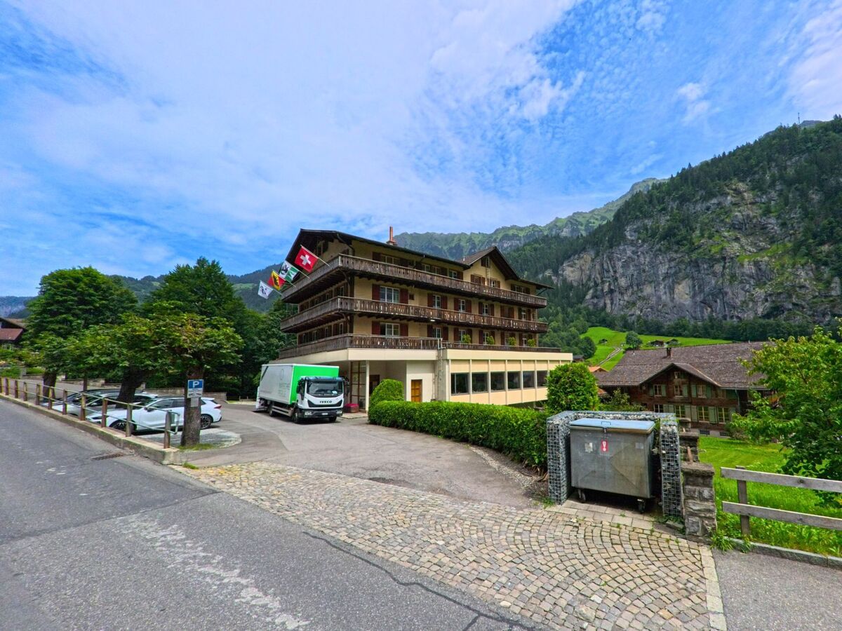 In Lauterbrunnen (3822, Kanton Bern) steht ein mehrstöckiges Gebäude mit Balkonen und wehenden Fahnen, im Hintergrund dominiert eine steile Felswand die Szenerie.