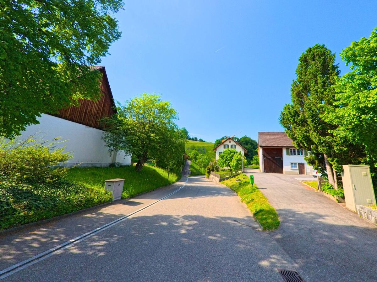 In Lauwil (4426, Kanton Baselland) führt eine Strasse bergauf an Wohngebäuden und Bäumen entlang, mit einer klaren Aussicht auf die umliegenden Hügel.