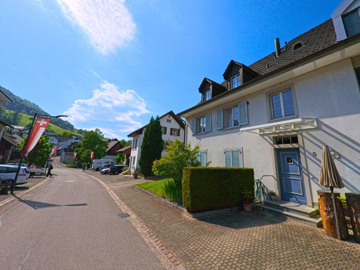 In Lauwil (4426, Kanton Baselland) führt eine Strasse an einem modernen Gebäude mit blauer Eingangstür und Schweizer Fahnen entlang, während der Blick auf die grünen Hügel im Hintergrund fällt.