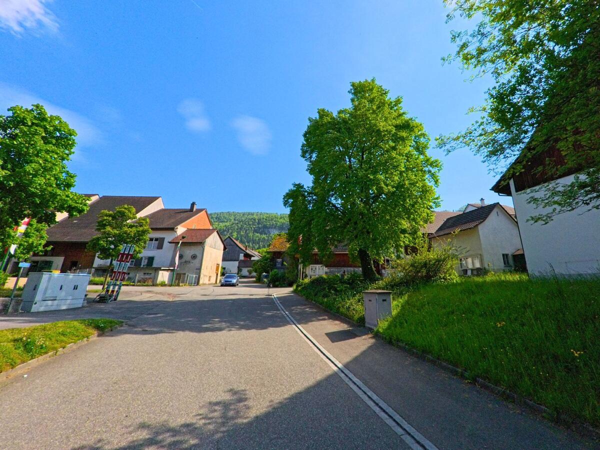 In Lauwil (4426, Kanton Baselland) verläuft eine Strasse an mehreren Gebäuden und Bäumen entlang, während die Hügel im Hintergrund sichtbar sind.