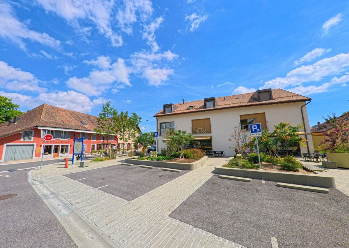 In Lavigny (1175, Kanton Waadt) grenzen ein weisses Wohnhaus mit Balkonen und ein rotes Gebäude mit Restaurant an einen Parkplatz mit Bäumen.