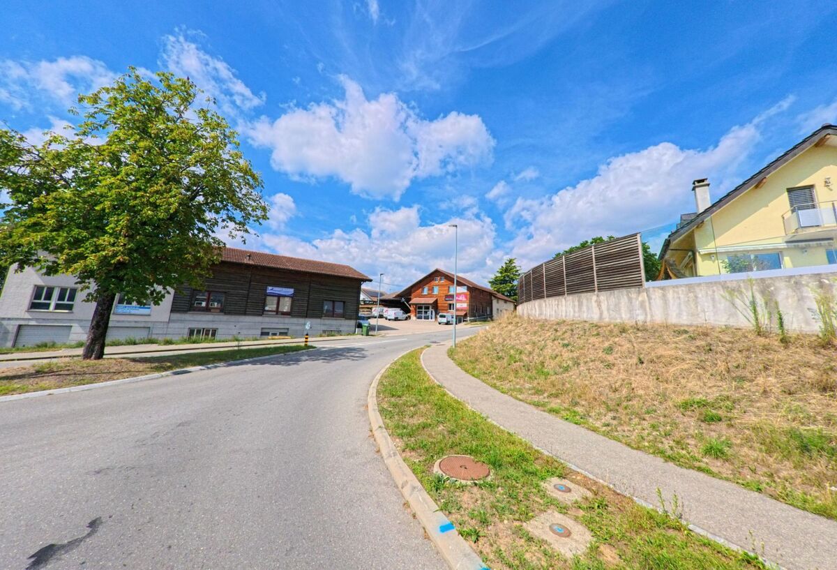 Eine leicht ansteigende Strasse in Lavigny (1175, Kanton Waadt) führt an Wohnhäusern und grossen Holzbauten vorbei, rechts verläuft ein Gehweg mit Böschung.