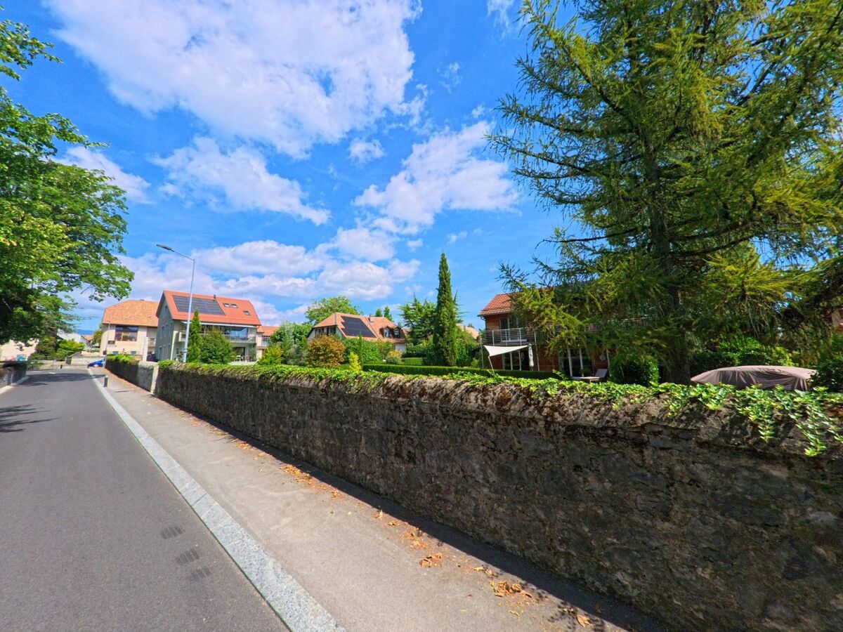Entlang einer Strasse in Lavigny (1175, Kanton Waadt) stehen moderne Wohnhäuser mit Solarpaneelen auf den Dächern, davor verläuft eine alte Steinmauer.