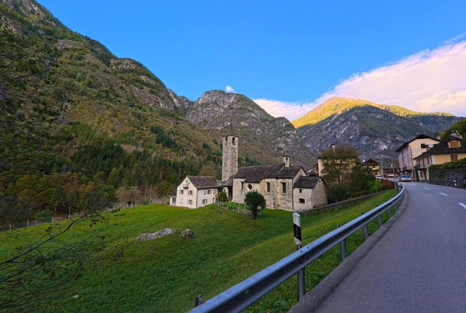 In Lavizzara (6694, Kanton Tessin) liegt am Rand der Strasse eine alte Steinkirche mit Turm, umgeben von grünen Wiesen und hohen Bergen im Hintergrund.