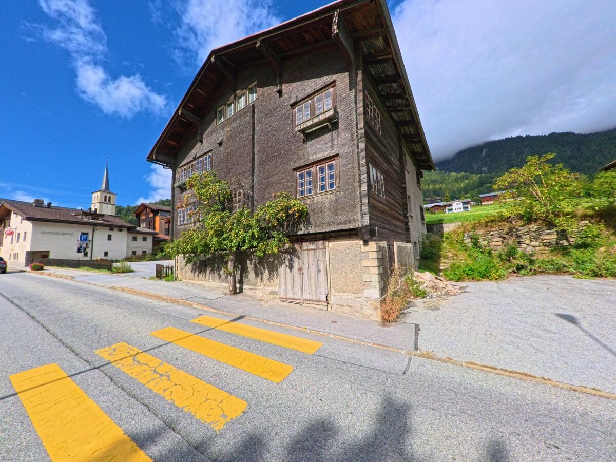 Ein altes Wohnhaus mit Holzfassade steht in Lax (3994, Kanton Wallis) an der Strasse, im Hintergrund sind die Kirche mit ihrem Turm und das Gasthaus Kreuz sichtbar.