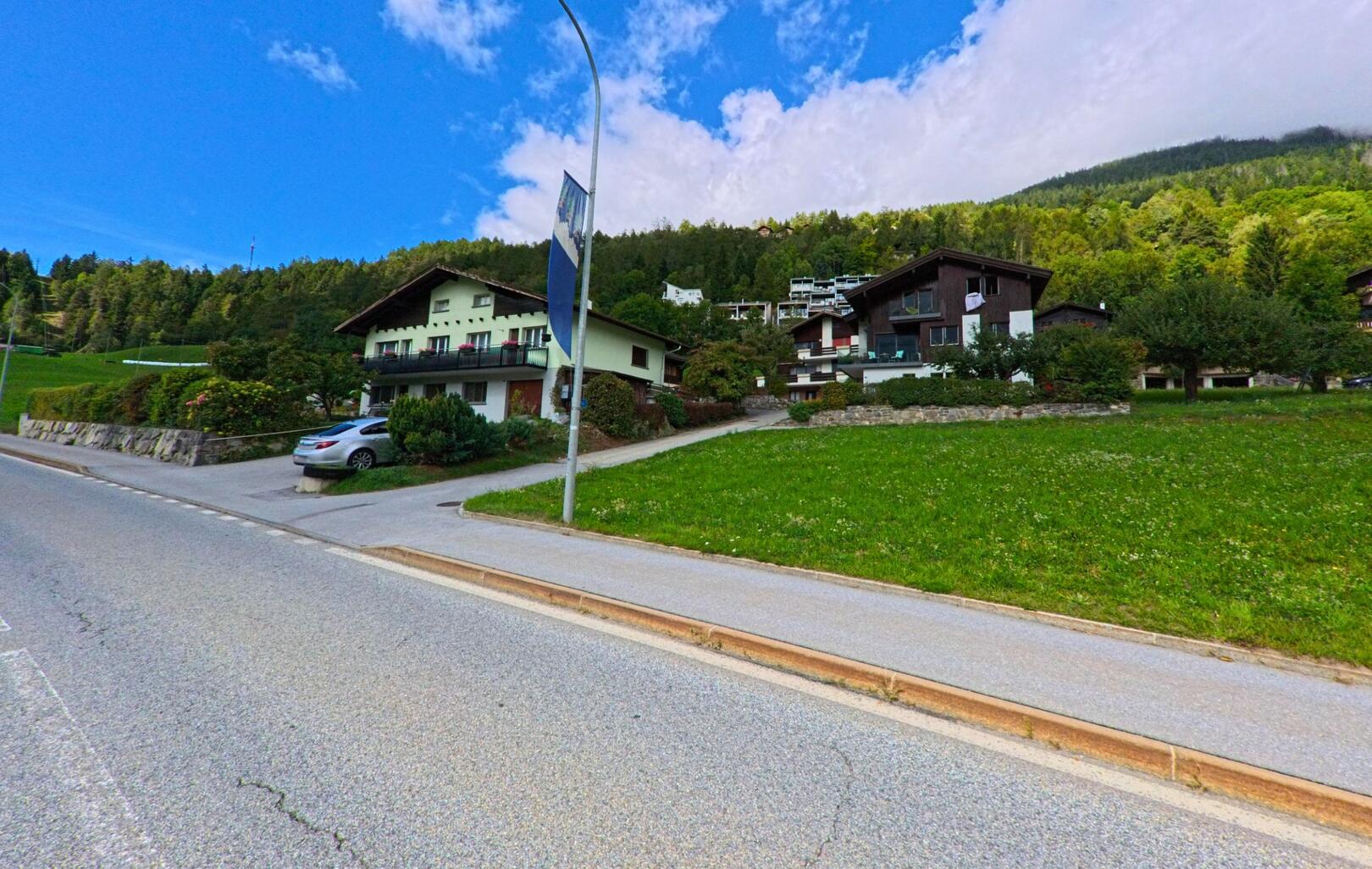 In Lax (3994, Kanton Wallis) stehen mehrere Einfamilienhaeuser mit Balkonen am Hang oberhalb der Hauptstrasse, eingerahmt von Wald und Wiesen.