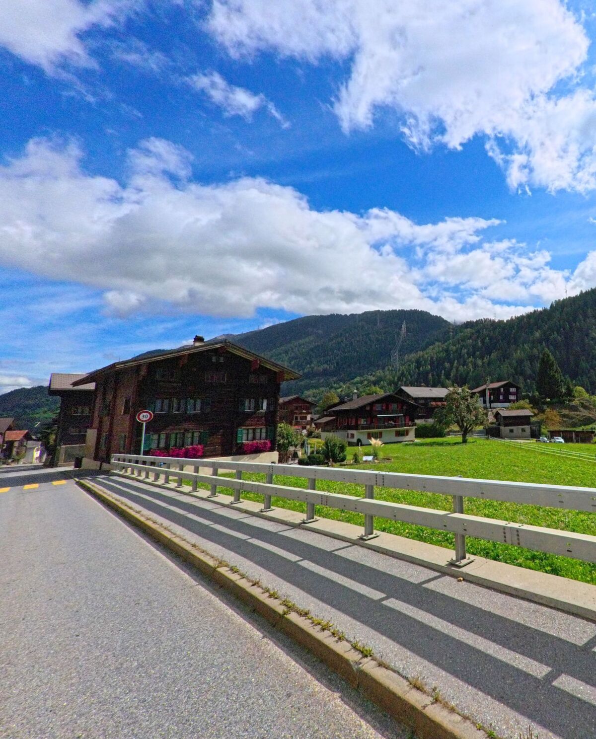 Am Ortsrand von Lax (3994, Kanton Wallis) stehen traditionelle Holzhäuser neben Feldern, im Hintergrund erhebt sich ein bewaldeter Berghang.