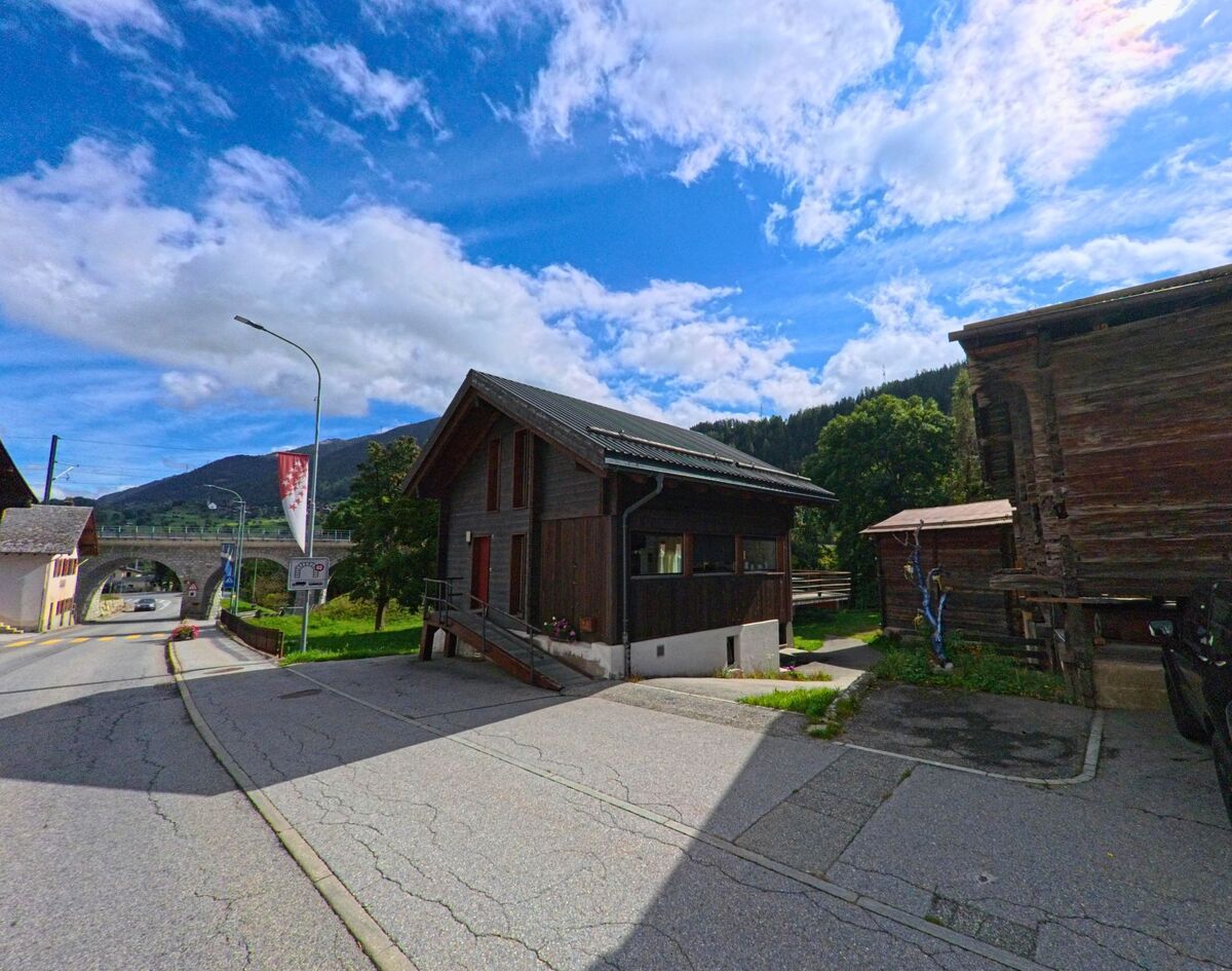 In Lax (3994, Kanton Wallis) steht ein modernes Holzhaus an einer schmalen Strasse, im Hintergrund ist das Steinviadukt der Bahnlinie sichtbar.