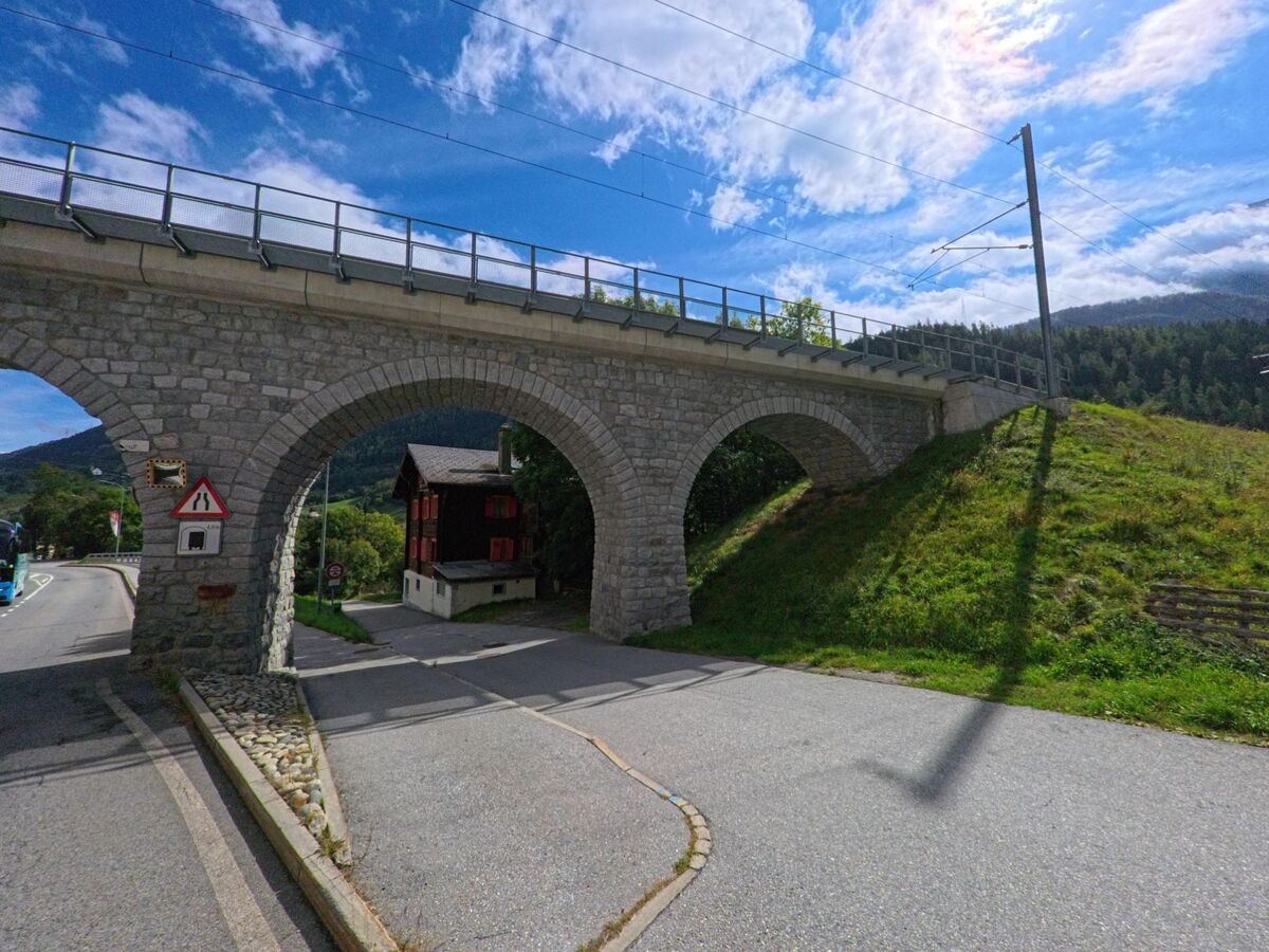 Ein Steinviadukt mit mehreren Bogen fuehrt die Bahnlinie in Lax (3994, Kanton Wallis) ueber eine Strasse, waehrend im Hintergrund Haenge und Waelder sichtbar sind.