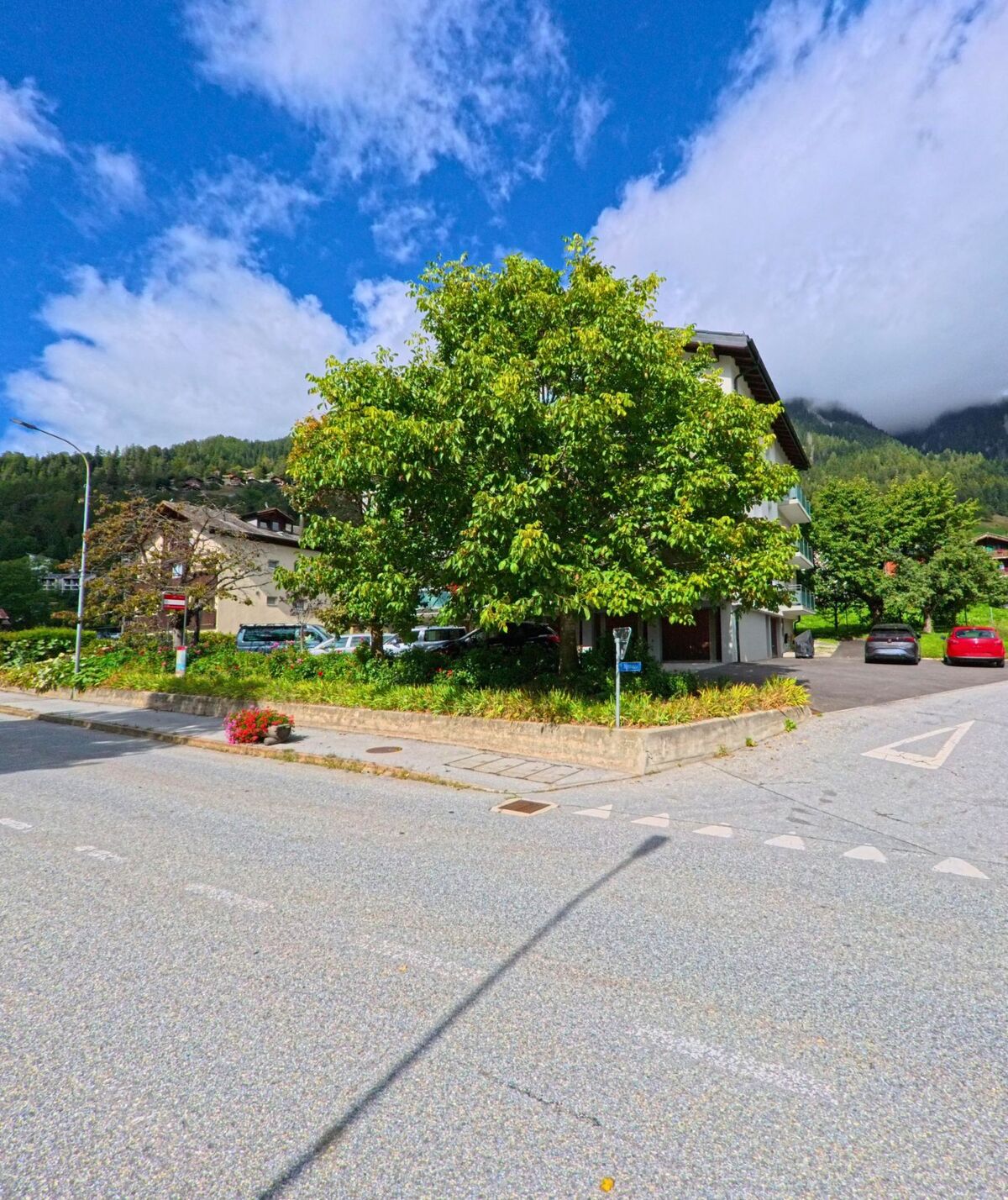 An einer Strassenecke in Lax (3994, Kanton Wallis) steht ein grosser Laubbaum vor einem Wohnhaus, umgeben von Blumenbeeten und Parkplaetzen.