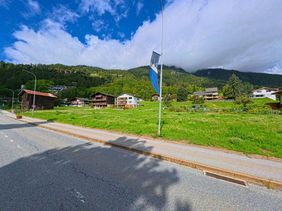 Von der Hauptstrasse in Lax (3994, Kanton Wallis) aus bietet sich der Blick auf Wohnhaeuser und Scheunen vor einem bewaldeten Berghang.