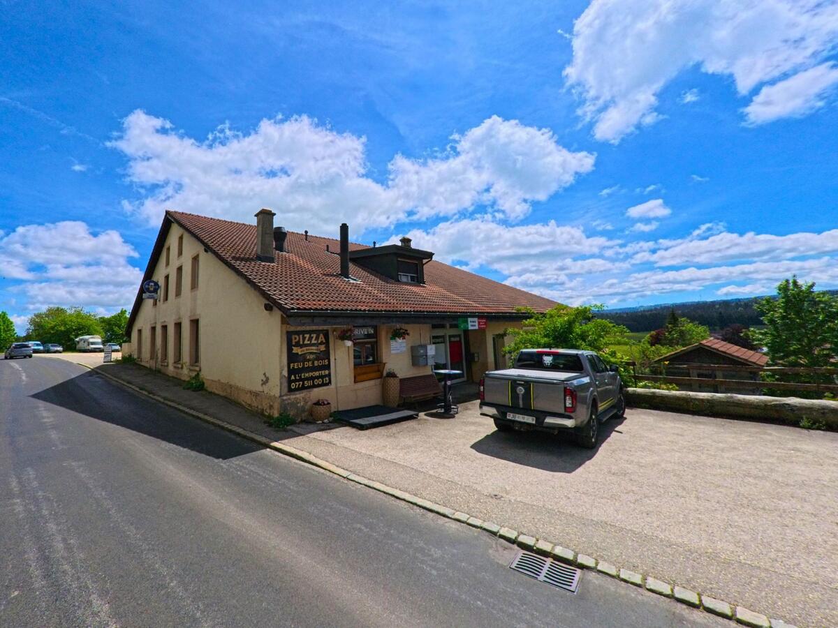 In Le Bémont (2360, Kanton Jura) ist ein Gebäude mit einem Pizzaladen und einer Terrasse zu sehen, daneben eine Straße mit parkenden Autos.