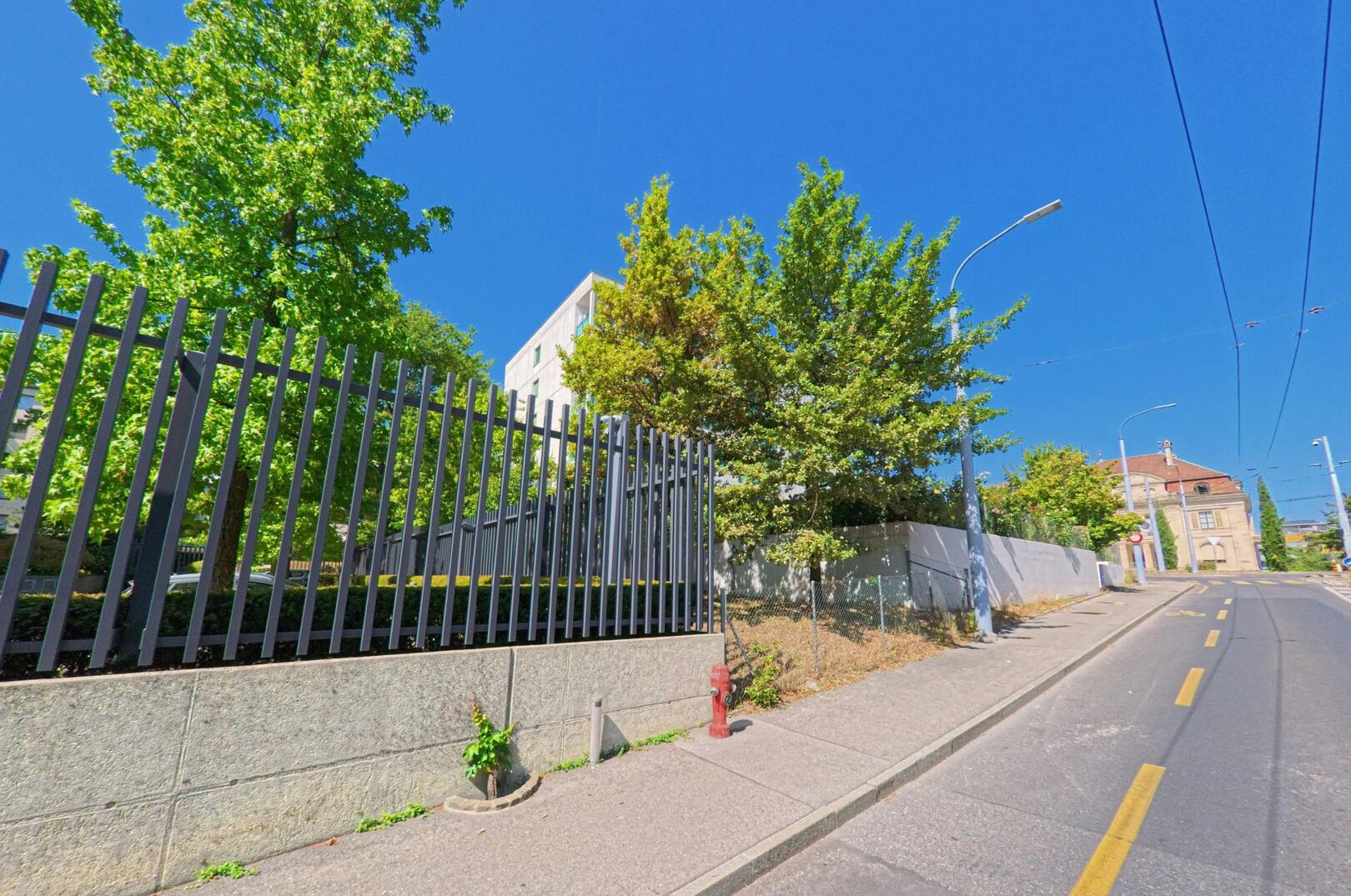 Eine ansteigende Strasse in Le Grand-Saconnex (1218, Kanton Genf) verläuft an einem hohen Metallzaun und grünen Bäumen vorbei, im Hintergrund ist ein älteres Gebäude zu sehen.