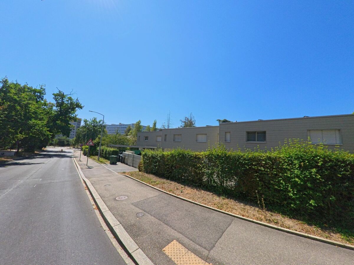 In Le Grand-Saconnex (1218, Kanton Genf) verläuft eine ruhige Strasse an niedrigen Wohnhäusern mit geschlossenen Fensterläden und grüner Hecke entlang.