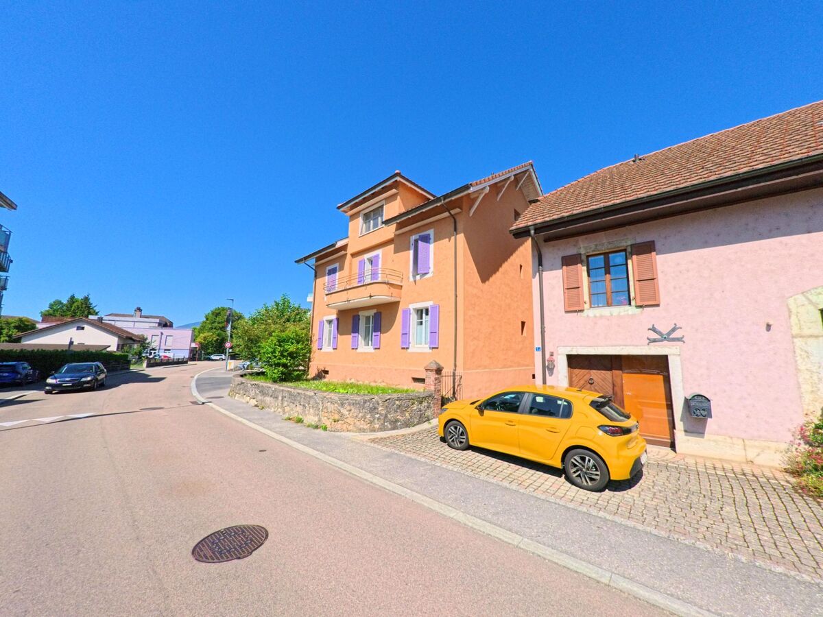 An einer leichten Kurve in Le Landeron (2525, Kanton Neuenburg) stehen farbige Wohnhaeuser, vor denen ein gelbes Auto parkt.