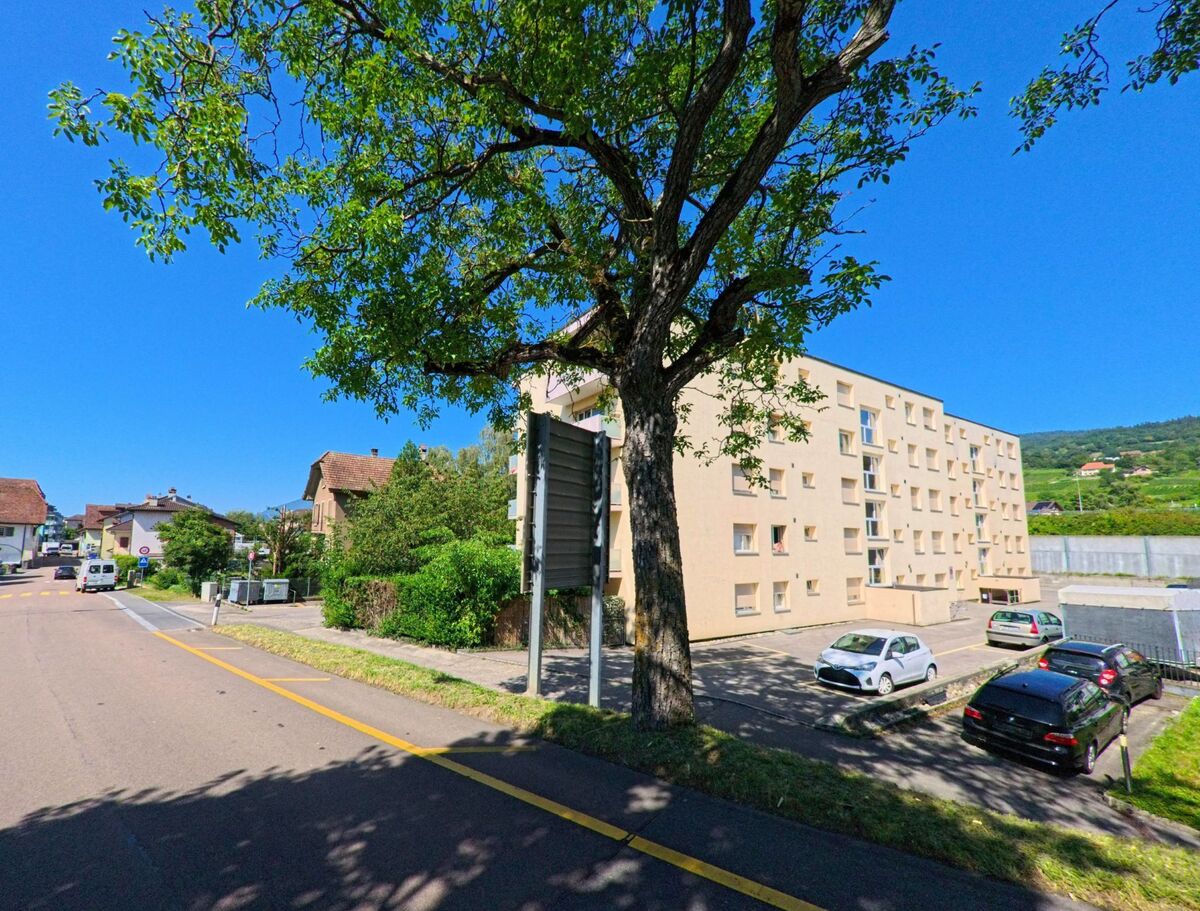 Ein mehrstöckiges Wohngebaeude mit kleinem Parkplatz steht in Le Landeron (2525, Kanton Neuenburg) an einer von Baeumen gesaeumten Strasse.