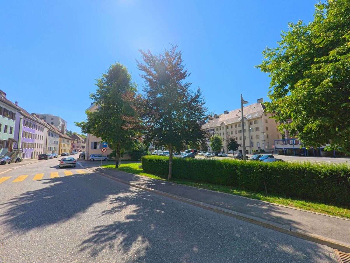In Le Locle (2400, Kanton Neuenburg) verlaeuft eine Strasse mit Fussgaengerueberweg entlang von Wohnhaeusern und gesaeumten Baeumen unter strahlender Sonne.