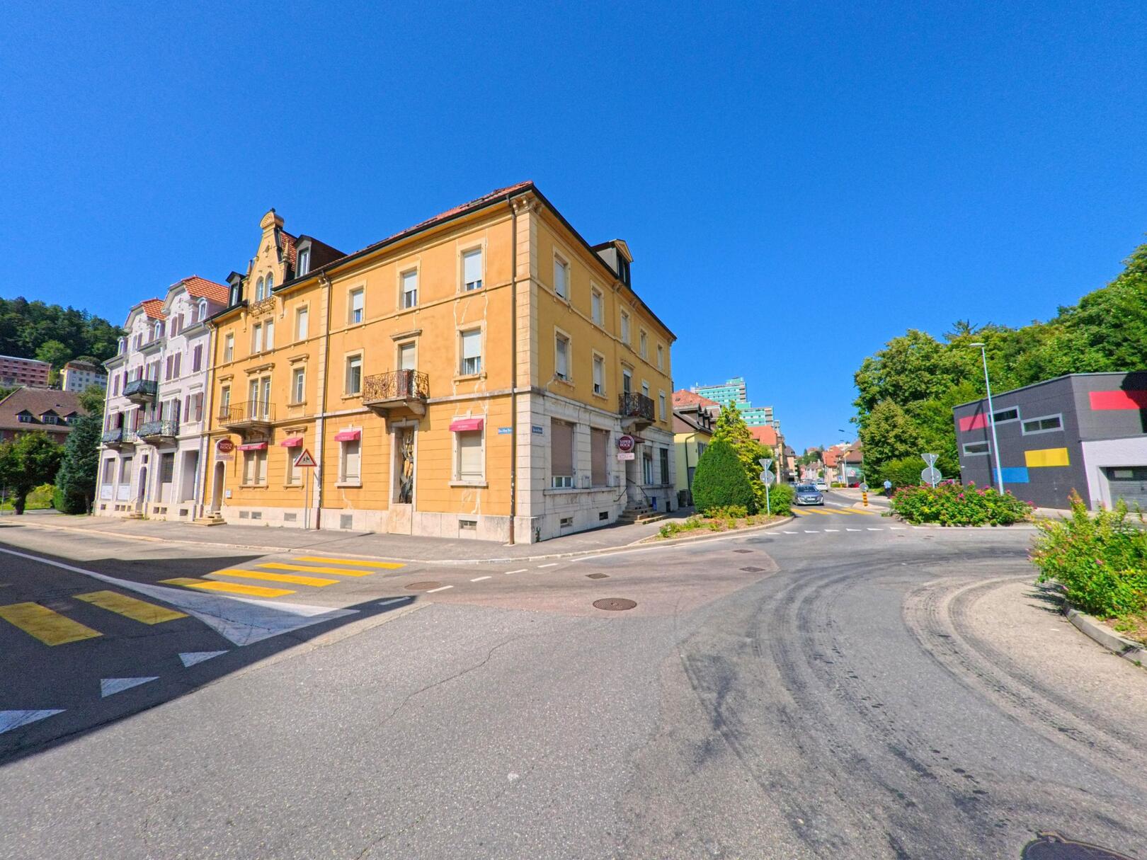 Mehrgeschossige Wohnhaeuser mit farbigen Fassaden saeumen eine Strassenkreuzung in Le Locle (2400, Kanton Neuenburg) bei sonnigem Wetter.