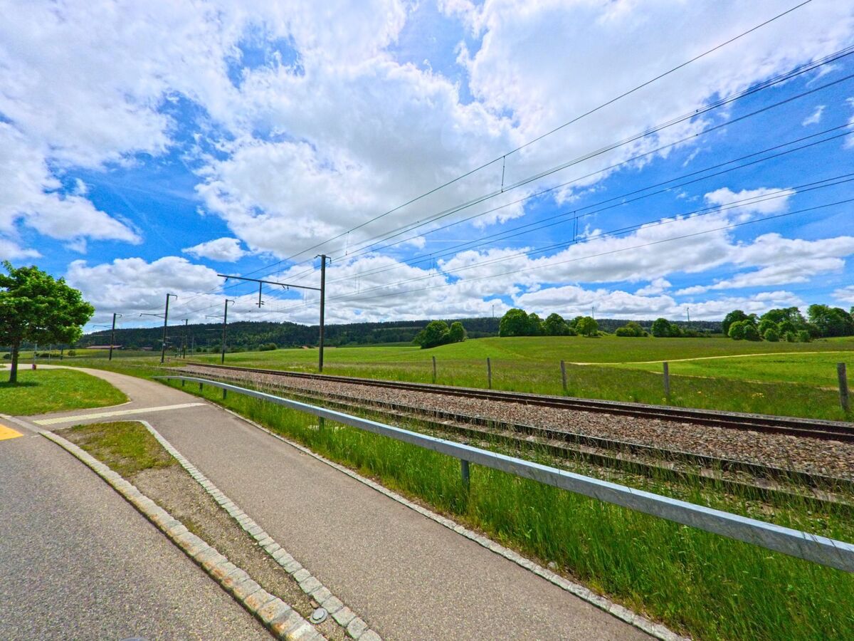 In Le Noirmont (2340, Kanton Jura) führt ein Gehweg entlang der Eisenbahngleise, umgeben von grünen Feldern und einem klaren Himmel.