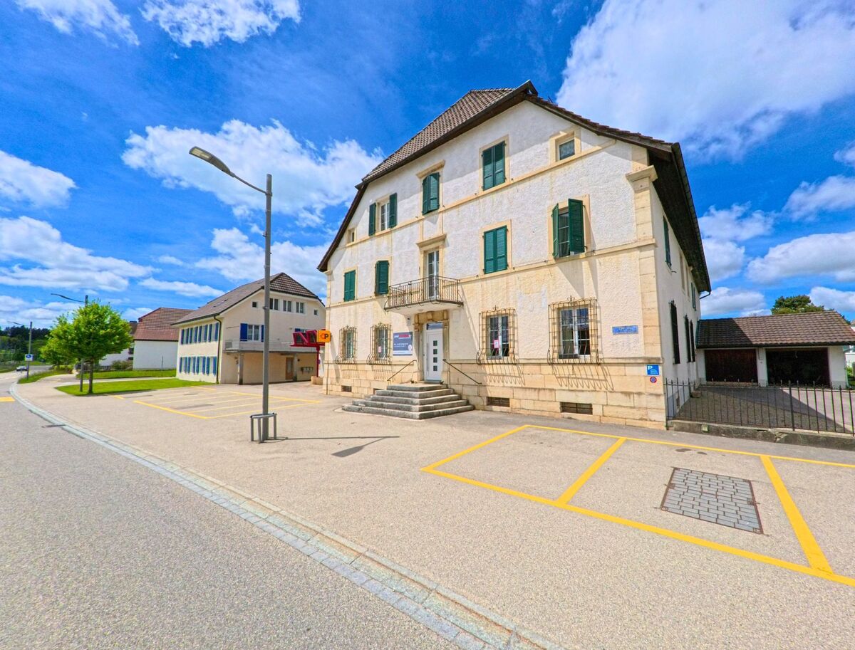 Ein historisches Gebäude in Le Noirmont (2340, Kanton Jura) mit grünen Fenstern und einer hohen Eingangstreppe, umgeben von einem Parkplatz und modernen Gebäuden.