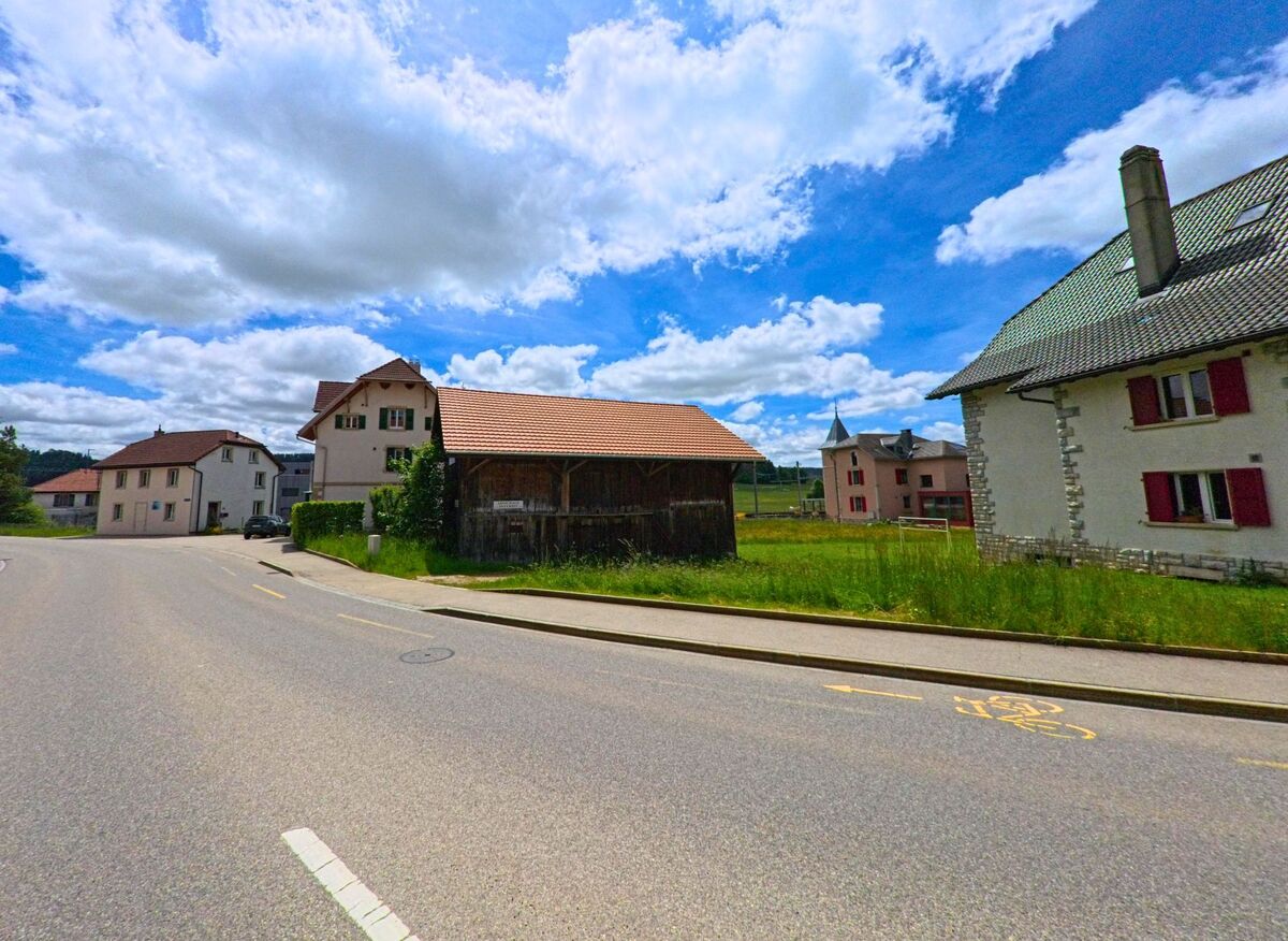 In Le Noirmont (2340, Kanton Jura) verläuft eine Straße an traditionellen Häusern und einem Nebengebäude vorbei, umgeben von grünen Wiesen und einem klaren Himmel.