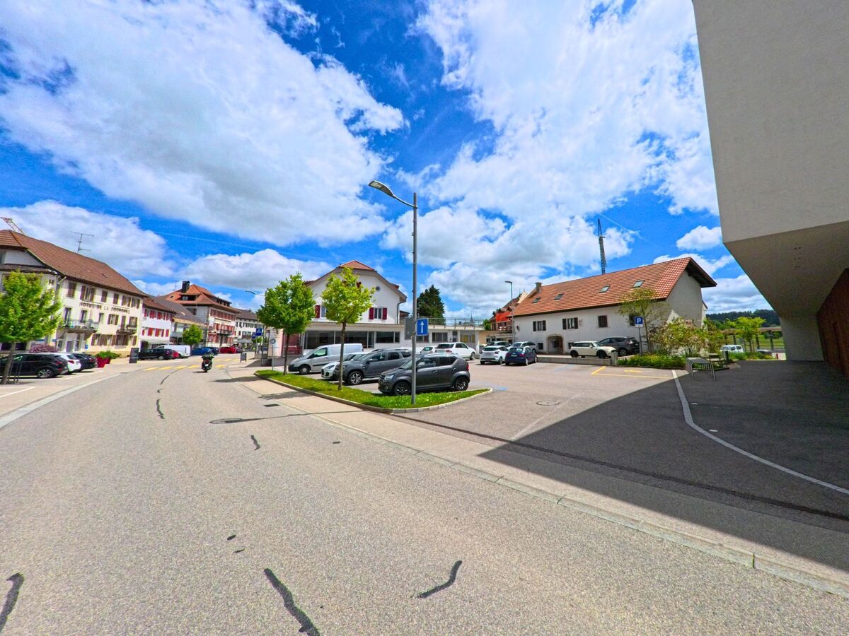In Le Noirmont (2340, Kanton Jura) verläuft eine Straße vorbei an modernen und traditionellen Gebäuden mit einem Parkplatz, umgeben von Bäumen und einem klaren blauen Himmel.