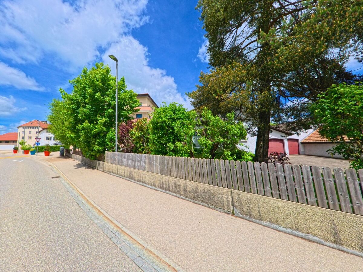 In Le Noirmont (2340, Kanton Jura) verläuft eine Straße mit einem Zaun, Bäumen und modernen Gebäuden, wobei bunte Pflanzgefäße die Szene verschönern.