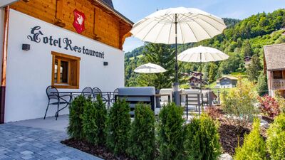 In Cresuz (1653, Kanton Freiburg) liegt das Hotel-Restaurant Le Vieux Chalet mit Sonnenterrasse, Sonnenschirmen und Blick auf die gruen bewaldeten Huegel.