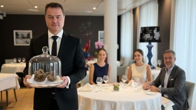 Im L’Ecu Vaudois in Begnins (1268, Kanton Waadt) praesentiert ein Kellner elegante Speisen, waehrend Gaeste in stilvollem Ambiente an einem festlich gedeckten Tisch sitzen.